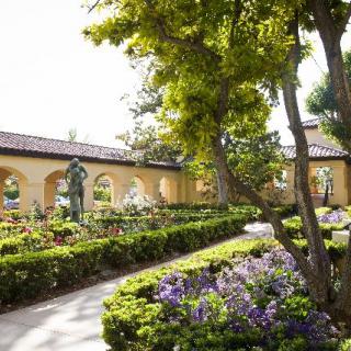 A tranquil garden with a pathway lined by blooming flowers and shaded by trees, alongside an arched wall.