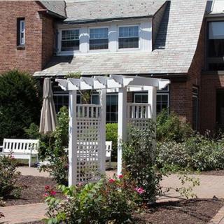 A charming garden area with a white pergola in front of a brick house, surrounded by benches and greenery.