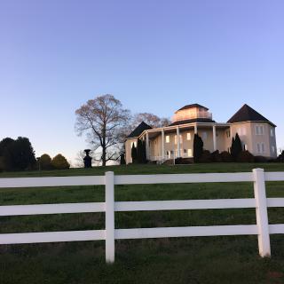 A rural house with a white fence and expansive green lawn under a clear sky.