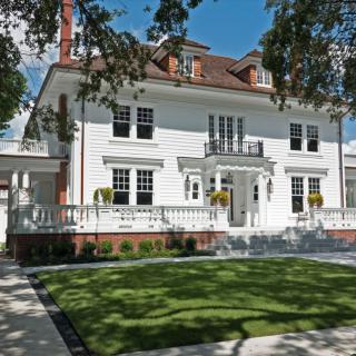 A large white colonial-style house with a manicured lawn and trees lining the property.