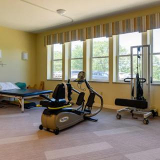 A rehabilitation room equipped with exercise machines and therapy tables, featuring large windows that allow natural light.