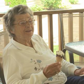 An elderly woman enjoys an ice cream cone while sitting on a porch.