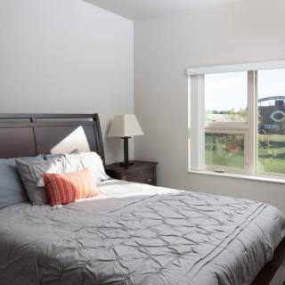 A cozy bedroom with a neatly made bed, featuring colorful pillows and a bedside lamp next to a window.