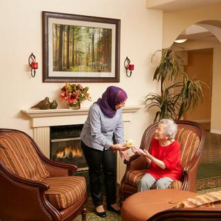 A caregiver assists an elderly woman in a cozy living room with a fireplace.