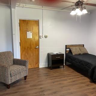A simple, sparsely furnished bedroom featuring a bed, chair, and wooden flooring.