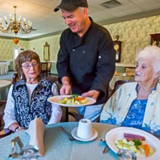 A chef serves a meal to two elderly women in a cozy dining setting.
