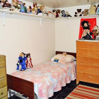 A cozy bedroom featuring a single bed with dolls and a dresser, adorned with numerous toys on shelves.