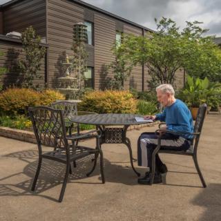An elderly person sitting at a table in a peaceful garden patio.