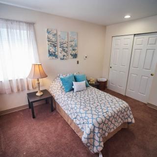 A cozy bedroom with a single bed, decorated with pillows and a bedside lamp, featuring a carpeted floor and a closet.
