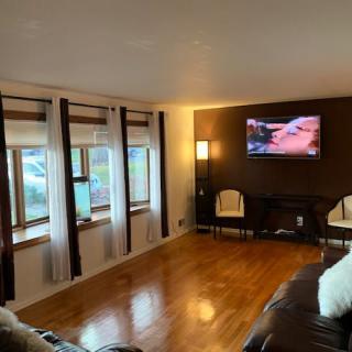A cozy living room with a TV mounted on the wall, wooden flooring, and several windows with curtains.