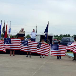 A group of individuals holding American flags during a speaking event, surrounded by various military flags.