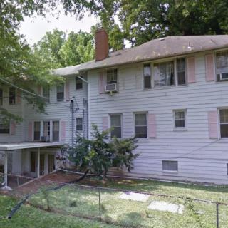 A large residential house with multiple windows and a chimney, surrounded by trees and a fenced backyard.