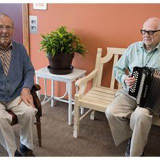 Two elderly men seated indoors, one playing an accordion.