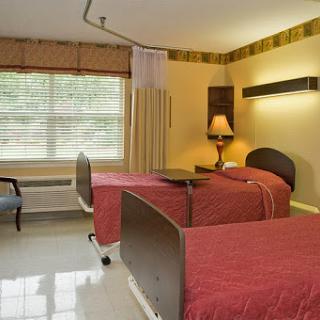 A cozy patient room featuring two beds, a lamp, and a large window for natural light.