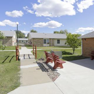 A peaceful residential area featuring benches along a pathway surrounded by green grass and buildings under a partly cloudy sky.
