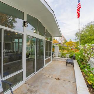 A modern building with large glass windows and a patio featuring outdoor seating, surrounded by greenery and an American flag in the background.