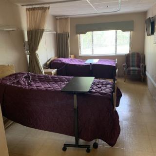 A hospital room featuring two beds with purple bedding, natural light coming through the window, and comfortable seating.
