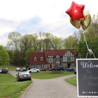 A charming house with a driveway adorned by balloons and a welcome sign, surrounded by lush trees.