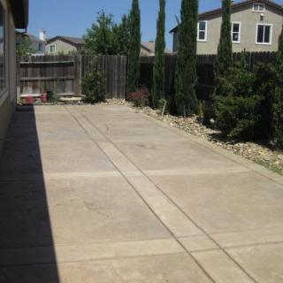 A concrete backyard space bordered by trees and a wooden fence, with houses in the background.