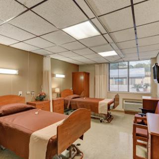 A view of a hospital patient room featuring two beds, essential furnishings, and natural light from a nearby window.
