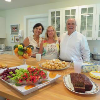 Three people in a kitchen enjoying a spread of fruits and baked goods.