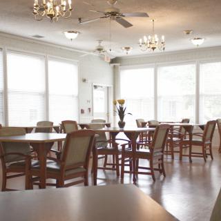 A bright and spacious dining room with multiple tables and chairs, illuminated by chandeliers and natural light from large windows.