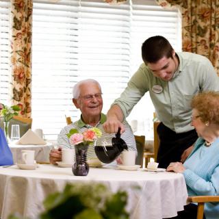 A caregiver is pouring coffee for elderly individuals sitting around a table in a cozy setting.