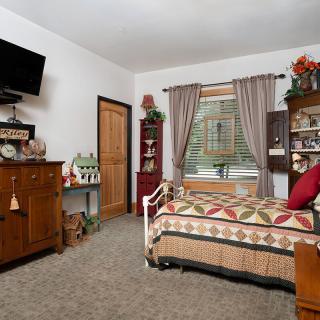 A cozy bedroom featuring traditional decor with a daybed adorned with a quilt, wooden furniture, and a wall-mounted TV.