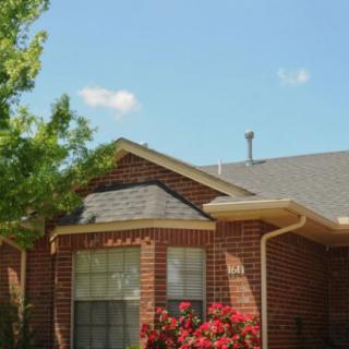 A brick house with a sloped roof, surrounded by trees and flowers under a clear blue sky.