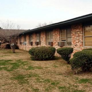 A single-story brick residential building with a grassy lawn and neatly trimmed bushes.