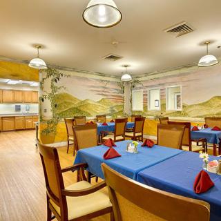 A cozy dining room with blue tablecloths and a mural on the wall, connected to a kitchen area.