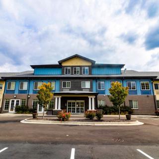 A modern commercial building with a parking lot in front, surrounded by small trees under a cloudy sky.