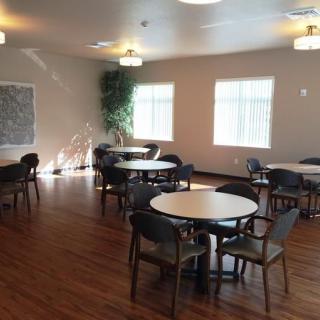 A spacious dining room with round tables and chairs on a wooden floor, lit by ceiling lights and natural light from the windows.