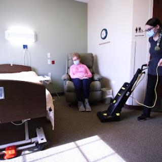 A healthcare worker cleans the hospital room while a patient sits in a chair.