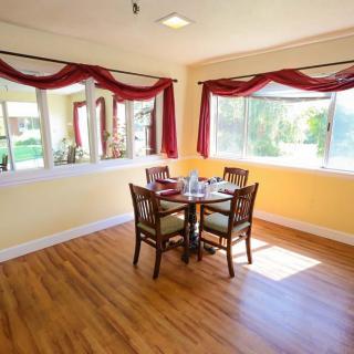 A cozy dining area with a wooden table and four chairs, surrounded by large windows with red curtains allowing natural light to fill the room.