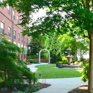 A serene garden pathway with lush greenery, leading to a fountain and surrounded by buildings.