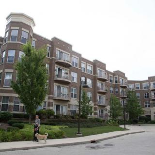 A view of a residential apartment building with landscaped surroundings and a person walking their dog on the sidewalk.