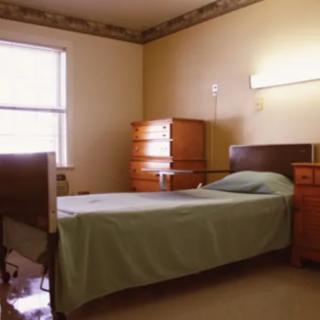 A minimalist hospital room featuring a single bed and wooden furniture, illuminated by natural daylight.