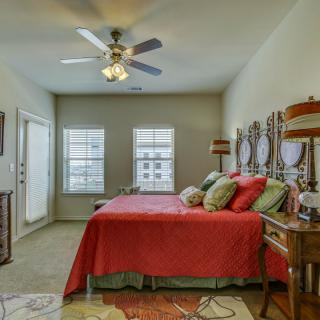 A cozy bedroom featuring a red bedspread, multiple pillows, and a ceiling fan with wooden blades. The room is well-lit by two large windows and is decorated with lamps and matching nightstands.