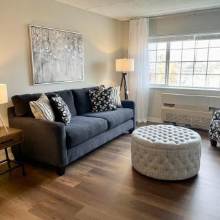 A modern living room featuring a dark sofa with patterned cushions, an ottoman, and floor lamps. The room is decorated with wall art and has a large window allowing natural light.