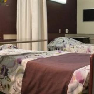 A hospital room with two beds, separated by a curtain, featuring floral sheets and a wall-mounted television.