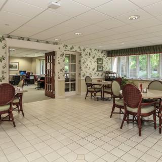 A cozy dining area with floral wallpaper and multiple tables and chairs, featuring large windows and a tiled floor.