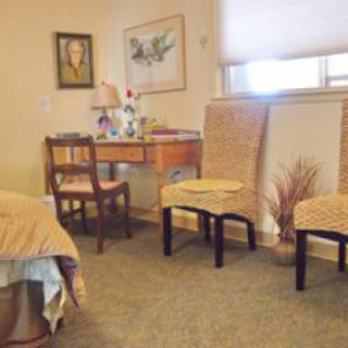 A cozy bedroom featuring a bed, two chairs, and a desk with decorative items and paintings on the walls.