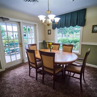 A cozy dining room with a round table and six chairs, illuminated by a chandelier, featuring carpeted flooring and large windows with curtains.