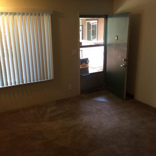 A small empty room with carpet flooring, a window with vertical blinds, and an open door leading outside.