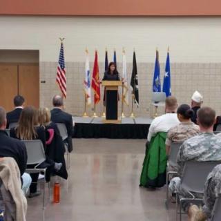 A speaker addresses an audience at an indoor event, with various flags displayed behind the podium.