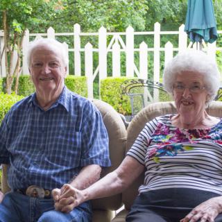An elderly couple sitting together outdoors, holding hands and enjoying the garden.