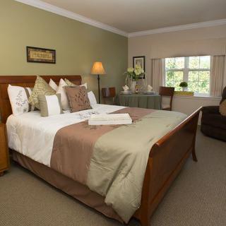 A well-decorated hotel room featuring a large bed with pillows and throws, bedside lamps, and a window overlooking greenery.