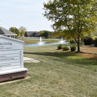 A serene outdoor setting featuring a sign, landscaped lawns, trees, a pond with fountains, and buildings in the background.