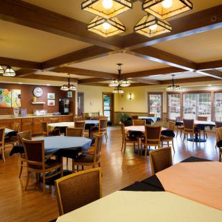 A spacious dining room with multiple round tables and chairs, featuring wooden flooring and ceiling lights.